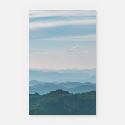 Eine ruhige Landschaft mit blau gefärbten Bergen, die sich in der Ferne unter einem weichen, teilweise bewölkten Himmel abzeichnen, mit grünen, bewaldeten Hügeln im Vordergrund.