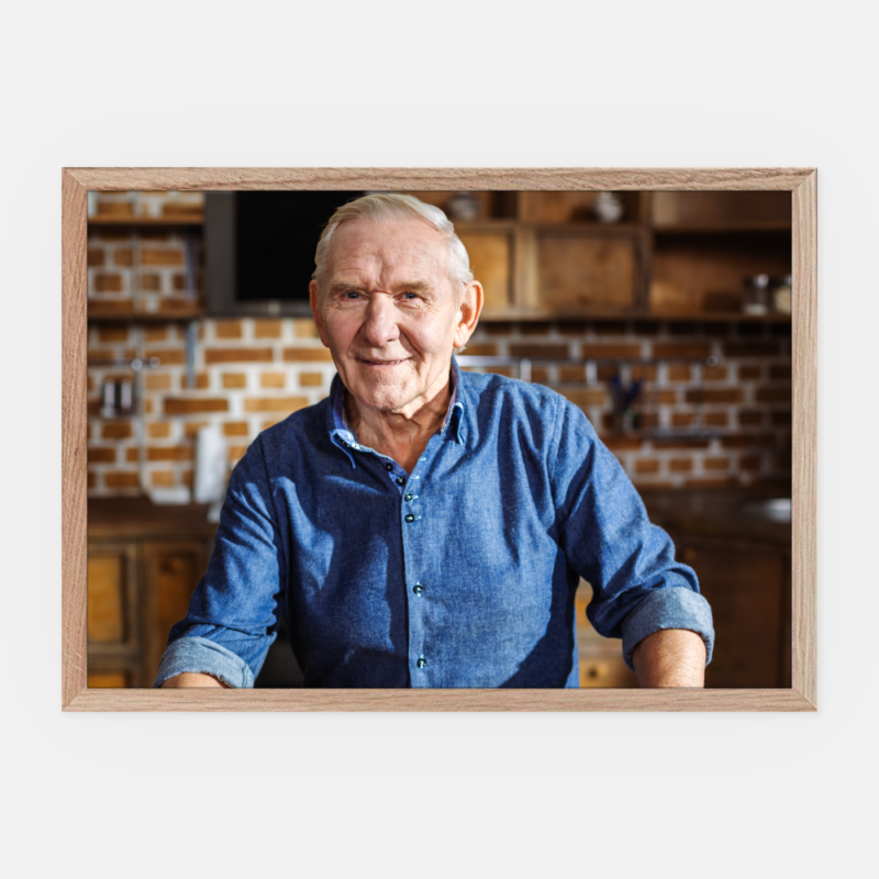 Ein lächelnder älterer Mann in einem blauen Jeanshemd steht in einer gemütlichen Küche mit Backsteinwänden und Holzschränken, eingerahmt von einem hellen Holzbilderrahmen.