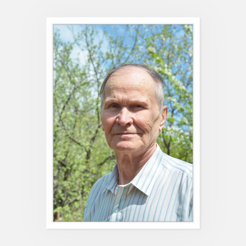 Älterer Mann mit kurzem grauem Haar und hell gestreiftem Hemd, steht draußen vor grünen Bäumen und blauem Himmel, blickt sanft in die Kamera - perfektes Portrait für Trauerfeier, um Erinnerung und Anteilnahme auszudrücken.