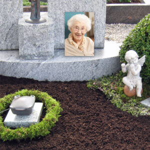 Ein Grabstein mit dem Foto einer älteren Frau, umgeben von Grünpflanzen. Eine kleine Engelsstatue befindet sich in der Nähe auf der rechten Seite des Grabes.
