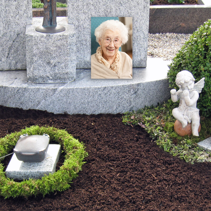 Ein Grabstein mit dem Foto einer älteren Frau, umgeben von Grünpflanzen. Eine kleine Engelsstatue befindet sich in der Nähe auf der rechten Seite des Grabes.