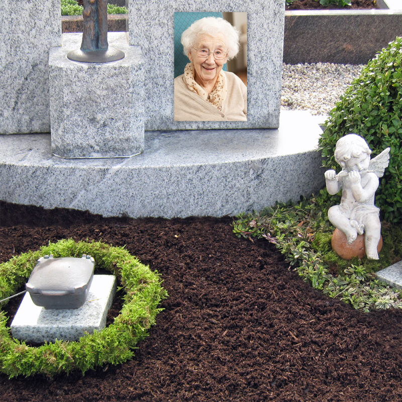 Ein Grabstein mit dem Foto einer älteren Frau ist auf einem Steinsockel aufgestellt. In der Nähe steht eine kleine weiße Engelsstatue inmitten von Grünzeug, und eine Kerze ruht in einem kreisförmigen Arrangement aus Grünpflanzen.